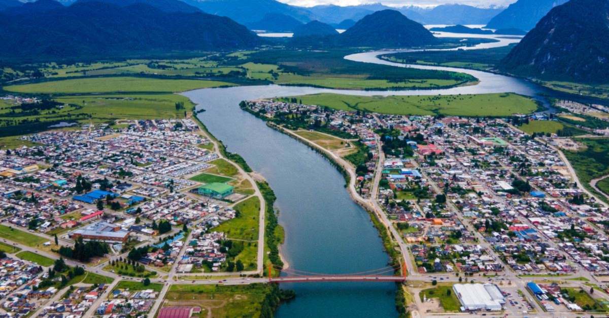Vista aérea de Puerto Aysén: el río cruzado por el puente Presidente Ibáñez, la ciudad en las dos orillas y los cerros del fiordo al fondo.